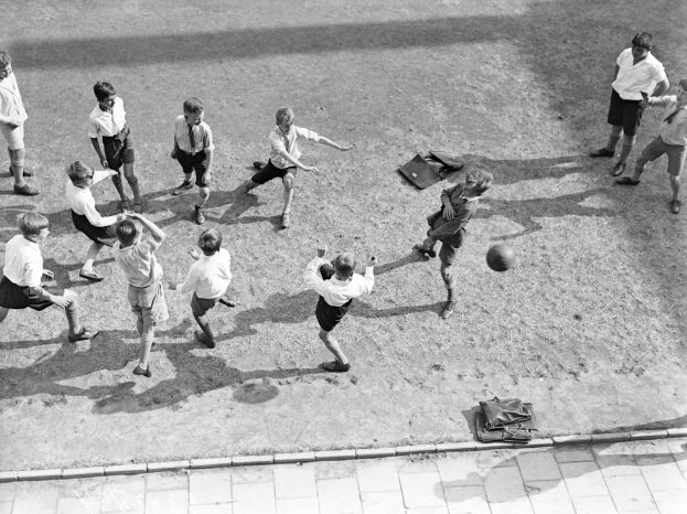 Eine Gruppe junger Jungs, die Fußball auf einem Feld in Schwarzweiß spielen, tragen Shorts und Schuhe und haben Taschen auf dem Boden um sie herum.