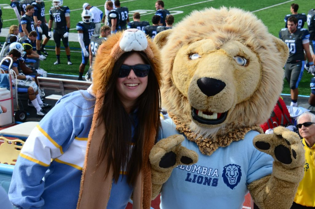 Eine Frau steht in der Nähe einer Person in einem Löwenkostüm, mit Spielern auf dem Boden dahinter und Zuschauern auf Bänken und Stühlen links.