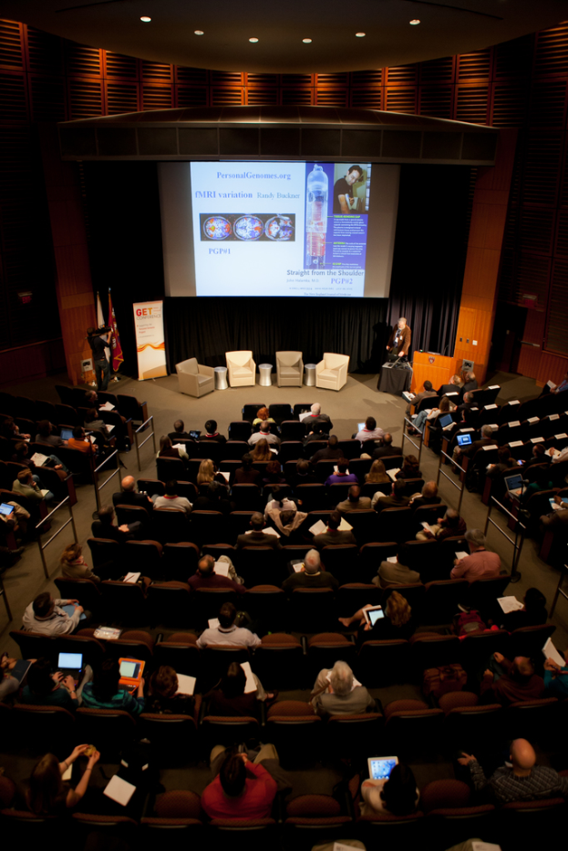 Ein mit sitzenden Menschen und Gegenständen gefüllter Auditoriumsraum, beleuchtet von vielen Lichtern, mit Fahnen links, einem Podium rechts und einem Werbeplakat links.