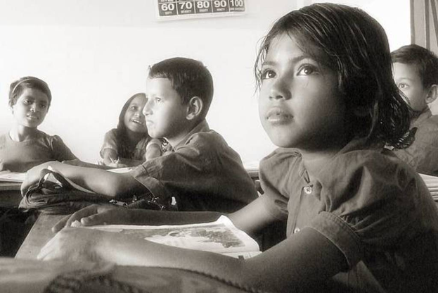 Eine Gruppe von Menschen sitzt an Tischen mit Büchern und Taschen, ein Kalender mit Zahlen und Text an der Wand und eine Tür oben rechts.