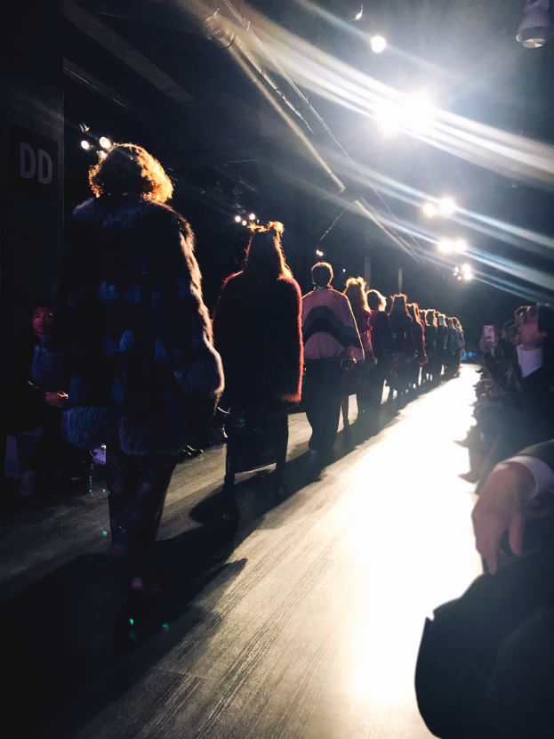 Eine Gruppe von Menschen läuft auf einem hell erleuchteten Laufsteg bei einer Fashion Show, mit Zuschauern auf beiden Seiten.