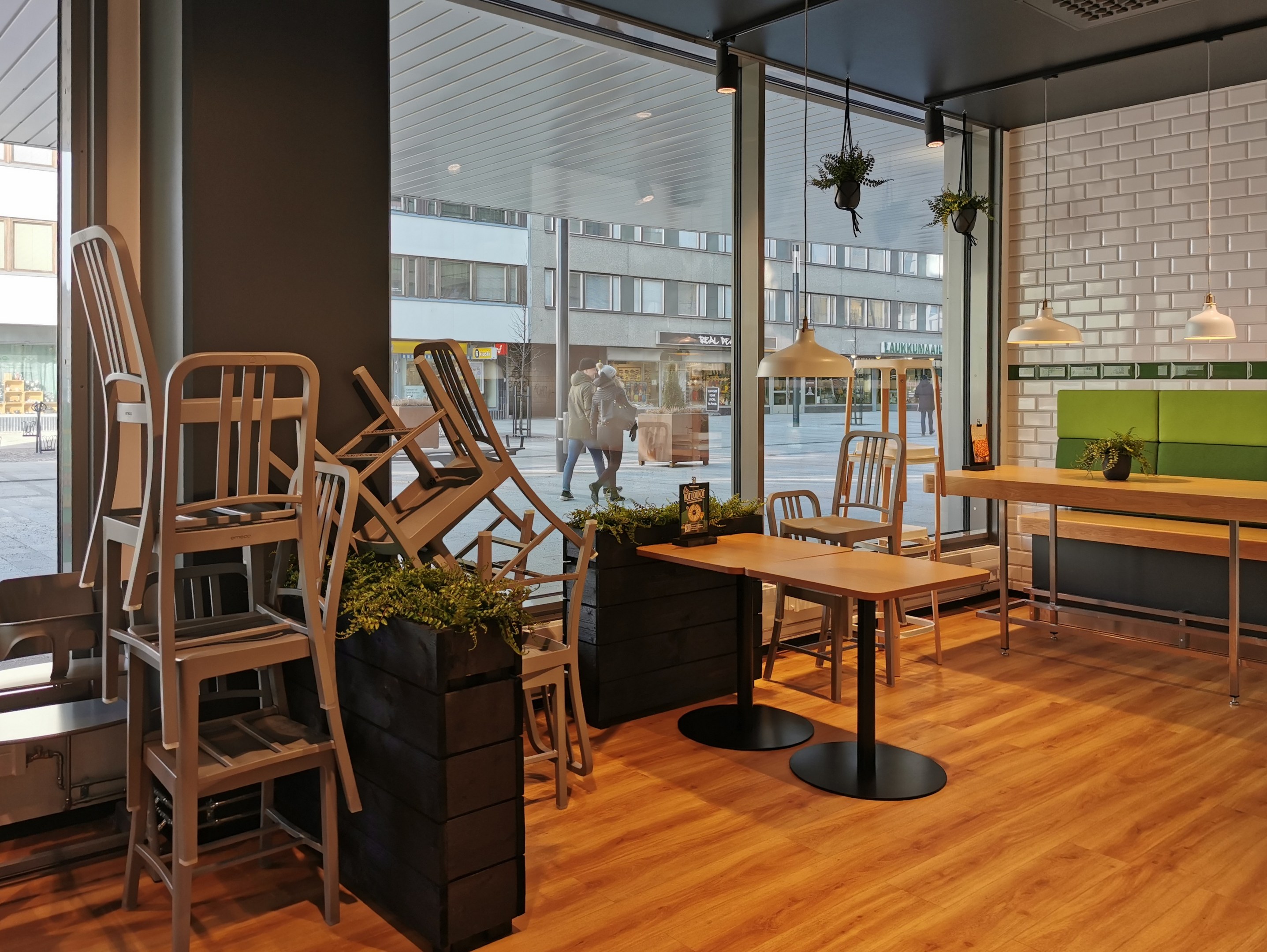 Ein Restaurant mit Holztischen und -stühlen drinnen, sichtbar durch ein großes Fenster, das Menschen auf einer Berliner Straße mit Gebäuden, Pfählen und Himmel zeigt.