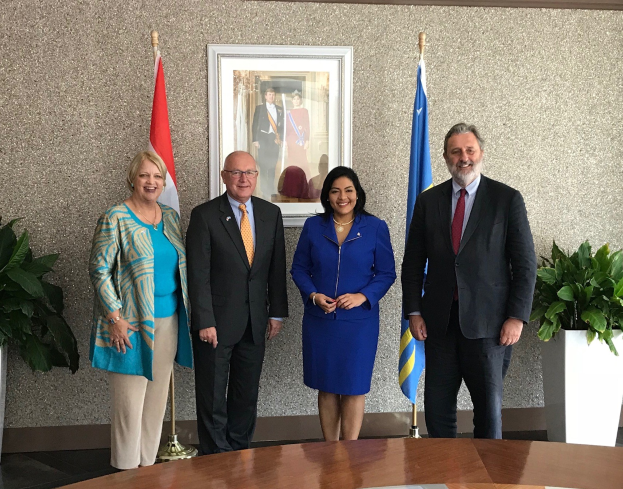 Gruppe von Menschen, die um einen Tisch mit Papieren stehen, umgeben von Fahnen, Topfpflanzen und einem Rahmenfoto an der Wand, was die Unterzeichnung eines U.S.-Canada Memorandum of Understanding anzeigt.