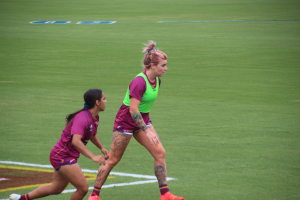 Zwei Frauen beim Fußballspielen auf einem Feld, eine in einem grünen T-Shirt und Shorts, die andere in einem pinken Kleid, beide mit Tattoos an Händen und Füßen.