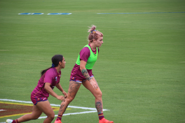 Zwei Frauen beim Fußballspielen auf einem Feld, eine in einem grünen T-Shirt und Shorts, die andere in einem pinken Kleid, beide mit Tattoos an Händen und Füßen.
