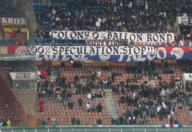 Ein volles Stadion mit Bannern und Stühlen im Vordergrund, einem Zaun unten und Treppen auf der linken Seite.