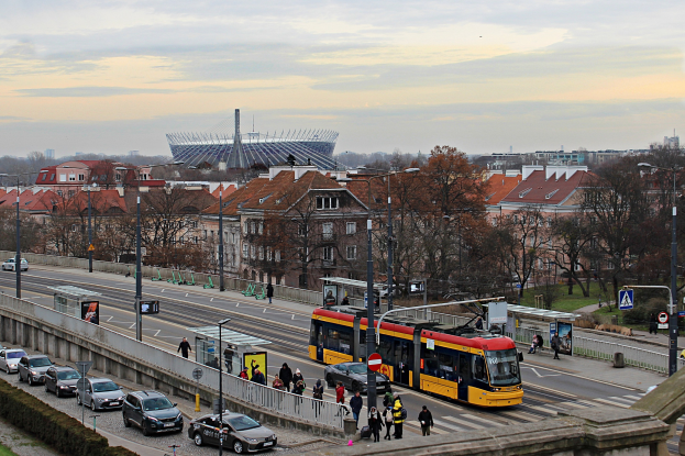 Eine gelbe und schwarze Straßenbahn fährt auf einer Stadtstraße mit hohen Gebäuden, umgeben von Autos, Fußgängern, Laternen, Schildern, Bäumen und Gras unter einem bewölkten Himmel.