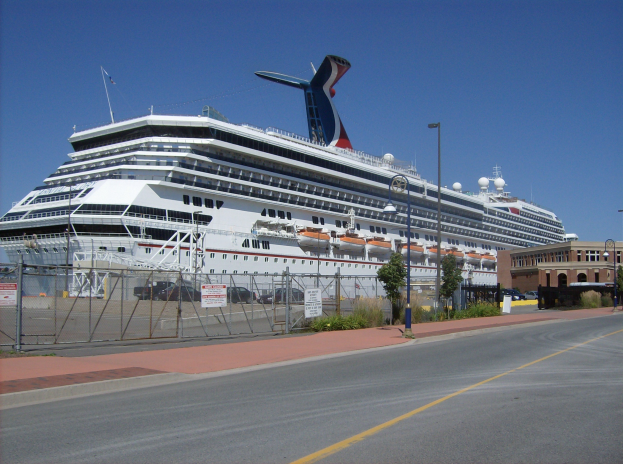 Großes Kreuzfahrtschiff liegt neben einer Straße, umgeben von einem Metallzaun, mit Bäumen, Schildern, Fahrzeugen, Strommasten und einem Gebäude im Hintergrund unter einem bewölkten Himmel.