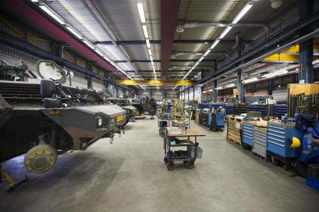 Großes Lagerinnere mit Maschinen, Fahrzeugen auf der linken Seite, Transportwagen und Holzboxen auf der rechten Seite sowie Deckenbeleuchtung.